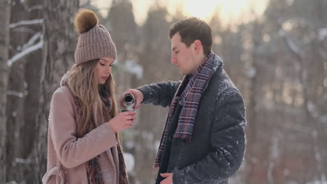 Couple-in-love-in-the-winter-forest-to-drink-tea-from-a-thermos.-Stylish-man-and-woman-in-a-coat-in-the-Park-in-winter-for-a-walk.